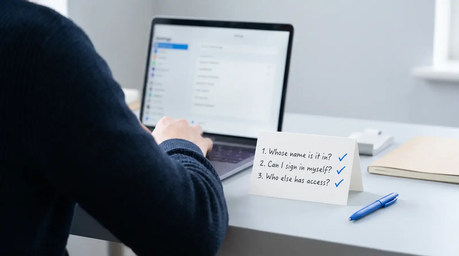 Business owner at a laptop with a desk card listing three questions: whose name is it in, can I sign in myself, who else has access